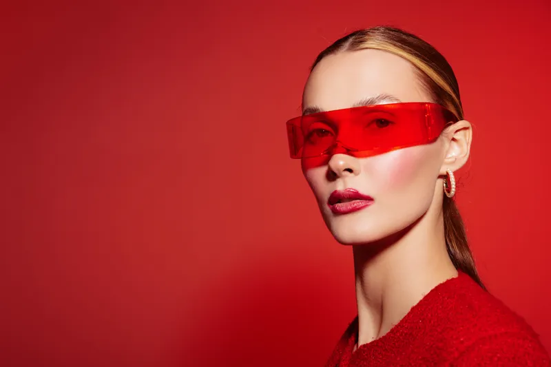Stylish woman with red sunglasses and red outfit poses against red background. Fashion portrait with bold makeup and sleek hairstyle. Concept of modern style and confidence.