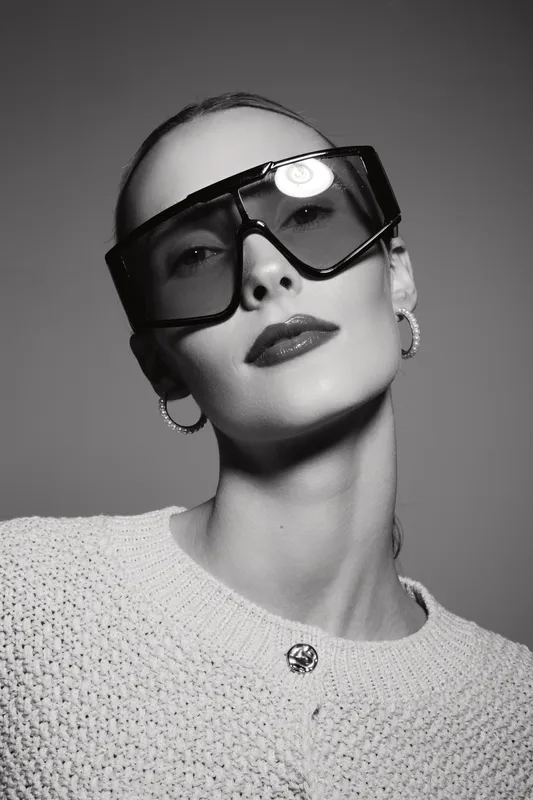 Stylish young white woman with pale skin wearing large sunglasses and hoop earrings looks confidently at camera. Minimalist black and white portrait with modern fashion concept.