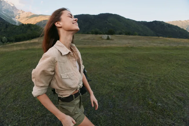 Young woman with long brown hair enjoys fresh air and freedom standing in an open grassy field with mountains in the background during daytime.