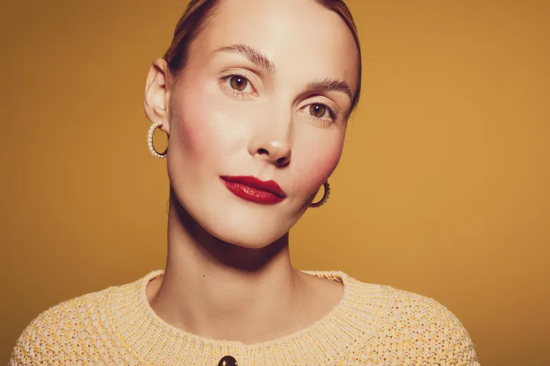 Portrait of a woman with red lipstick and gold hoop earrings wearing a beige sweater. Warm studio lighting highlights her clear skin and calm expression on a yellow background.