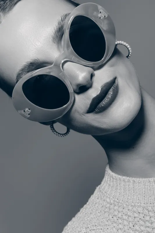 Close-up of a white woman with pale skin wearing round sunglasses and hoop earrings in a studio setting with soft lighting.