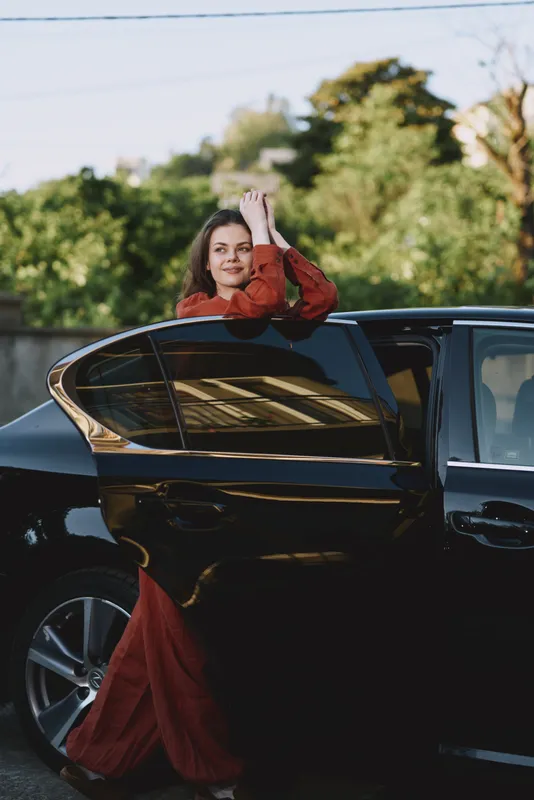 Cheerful woman outdoors, leaning from a sleek car window, red dress flowing, sunlit moment, relaxed posture, natural colors, lush greenery background, candid lifestyle vibe