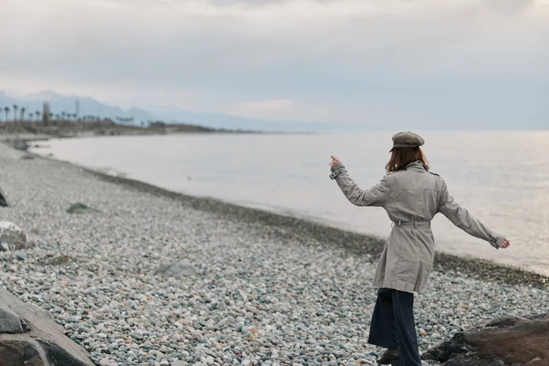 A woman in a coat and hat walks along a rocky beach near the ocean on a cloudy day. The scene conveys solitude, nature, and cool weather by the sea shore.