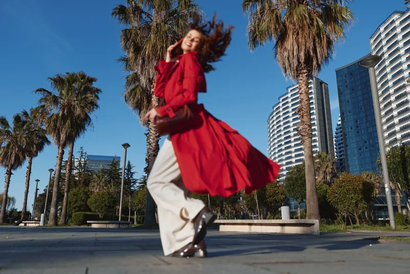 Cheerful woman in a red coat twirls outdoors in bright sunlight, urban backdrop with palm trees and tall buildings, dynamic motion and vibrant colors with breezy motion.