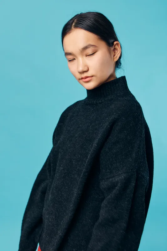 Young woman with black hair wearing oversized black sweater stands with eyes closed on blue background. Calm expression, casual clothing and simple hairstyle in studio portrait.