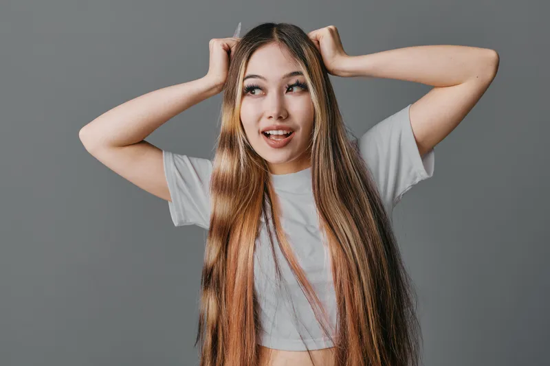 Young woman with long hair wearing casual shirt posing against gray background with playful expression and hands on head for lifestyle, emotion and portrait photography.