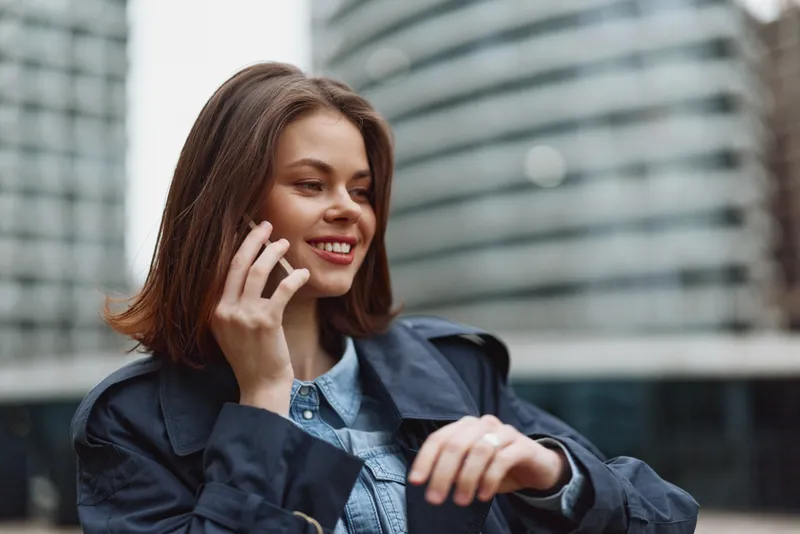 A smiling woman speaks on a smartphone in an urban outdoor setting, wearing a denim jacket and casual blazer. Contemporary cityscape conveys connectivity and everyday communication.