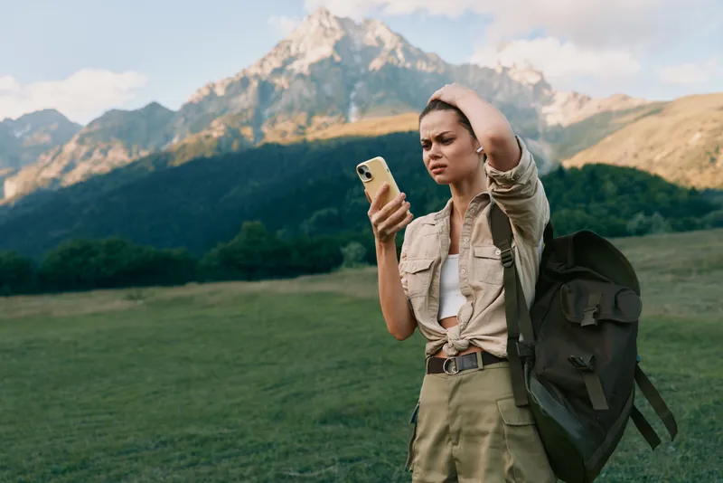 A woman outdoors checks her phone while wearing a hiking backpack, framed by rugged mountains, capturing a spontaneous travel lifestyle moment on a bright day with open fields.