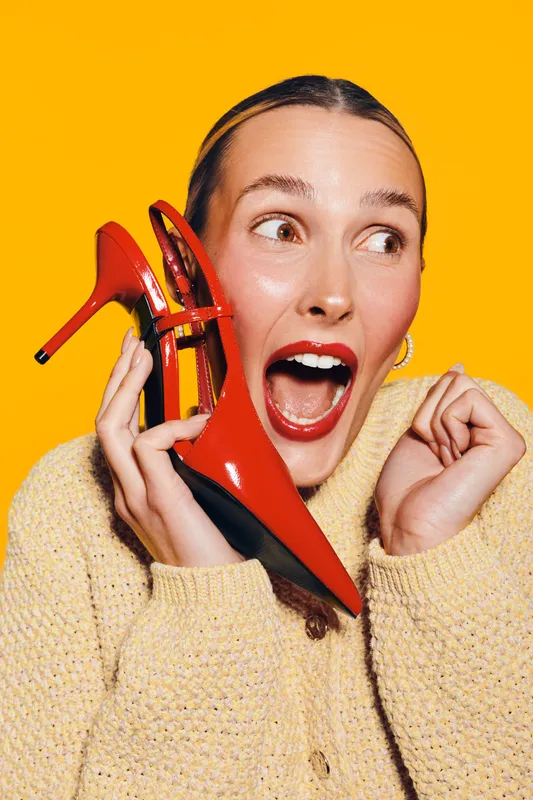 Excited woman in beige sweater holds red high heel shoe near face, mouth open in surprise. Bright yellow background highlights fashion and joyful expression concept.
