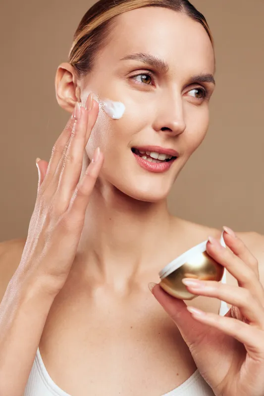 Young woman applying face cream on cheek with fingers, smiling softly on beige background, concept of skincare, beauty routine, hydration, and self-care with natural lighting.