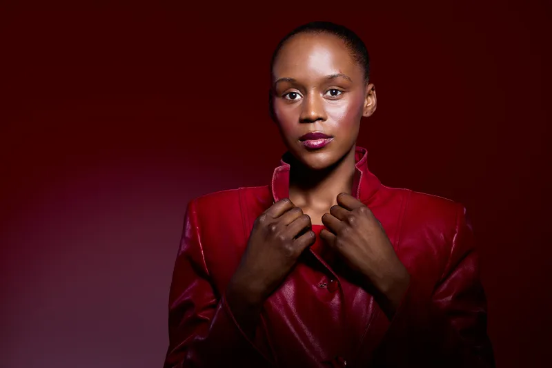 Confident African woman with dark skin wearing a red jacket poses against a dark red background. Studio portrait with dramatic lighting emphasizes strength and elegance.