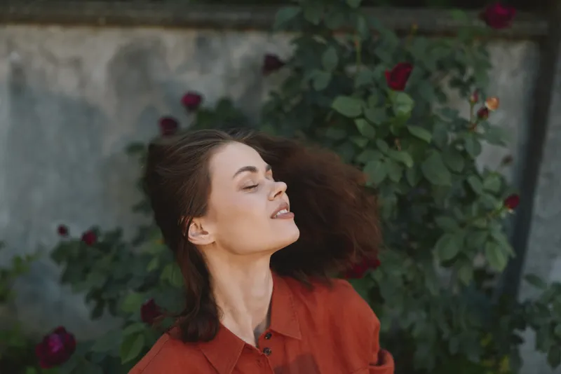 Young woman in an orange shirt enjoys a breezy moment among blooming roses, eyes closed and smiling, as hair dances in the garden courtyard, sunlit