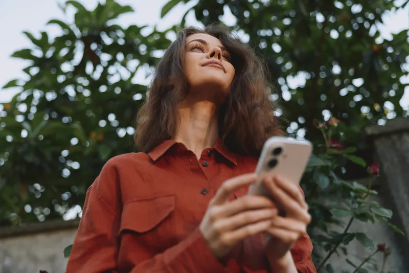 A woman stands outdoors in a red shirt, looking up while holding a smartphone. Bright natural light and lush foliage frame a focused, casual lifestyle moment for everyday creativity