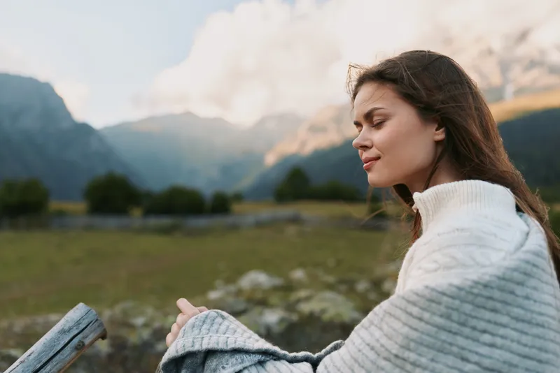 Calm woman in a cozy sweater outdoors on a mountainside, embracing nature and lifestyle moments in a bright, reflective mood.