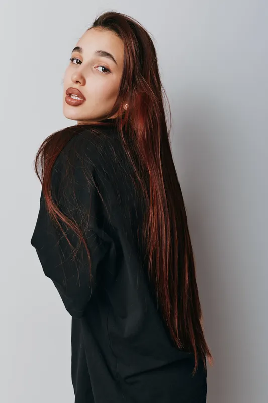 Young woman with long brown hair wearing black sweater posing and looking over shoulder against white background in studio. Female model with natural makeup and casual style portrait.