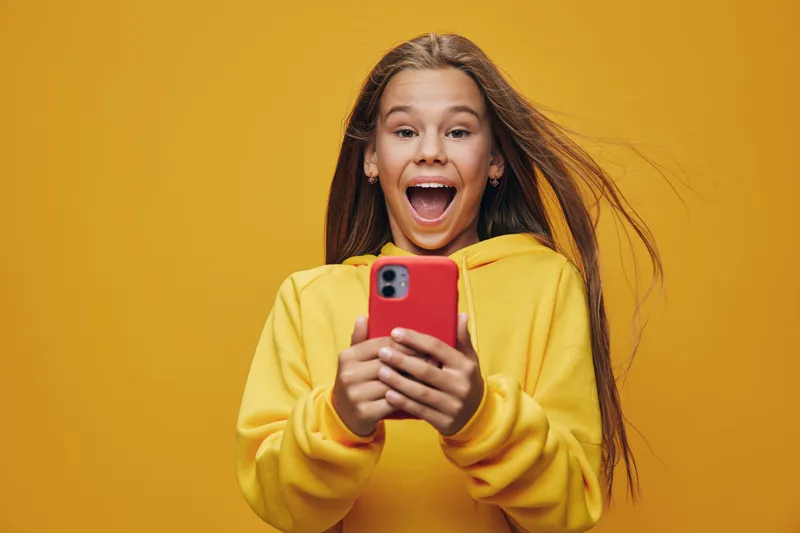 Girl wearing yellow hoodie holds red smartphone with wide smile on orange background. Happy teenager with long hair using mobile phone for social media or messaging.