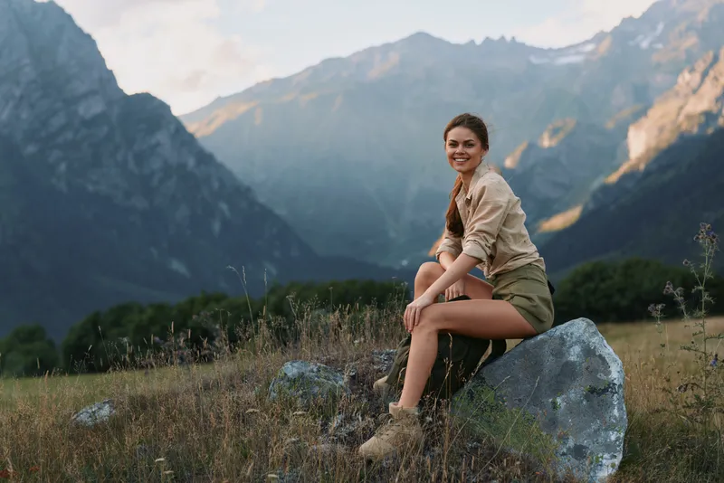 A young woman sits on a rock in a mountain meadow, smiling softly as she enjoys a peaceful, outdoor escape with rugged peaks in the distance.