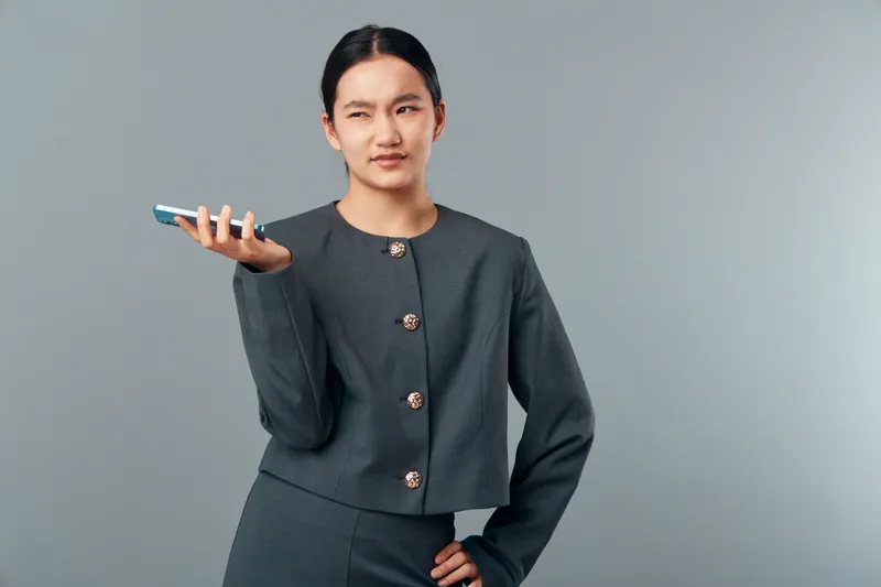 Woman in gray suit holding smartphone with thumb up expressing skepticism or doubt standing against gray background. Professional female with hand on hip and confused face gesture and look.