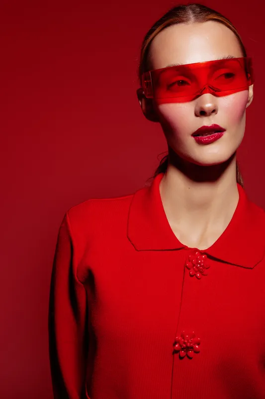 Fashion model in red sweater and red translucent visor poses against red background. Concept of modern style, bold colors, and futuristic accessories in studio lighting.