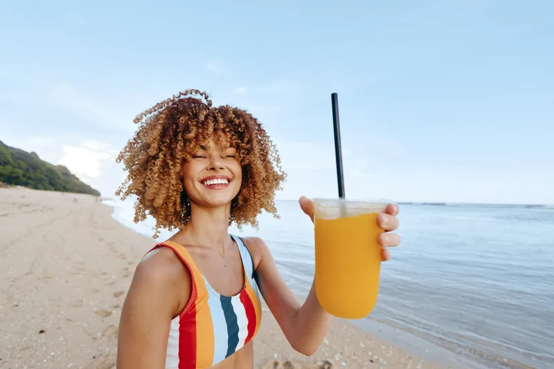 Smiling woman with curly hair on a sunny beach, holding a yellow drink with straw, summer vacation feeling, ocean horizon and waves, relaxed pose in natural light