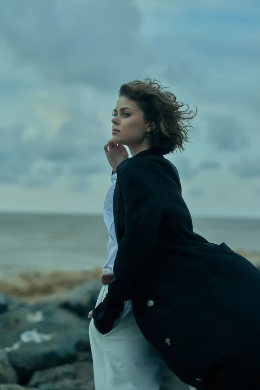 Wind, sea, coat, fashion, outdoors, woman, ocean, clouds, dramatic mood, windswept figure stands by the shoreline, gazing toward the horizon, capturing a stylish, cinematic seaside moment