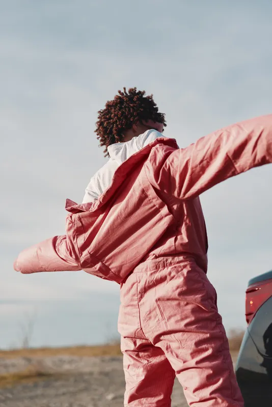 Outdoor person in a pink jumpsuit stands with arms wide, capturing motion, energy, and joy as they celebrate a moment near a vehicle in an open landscape.