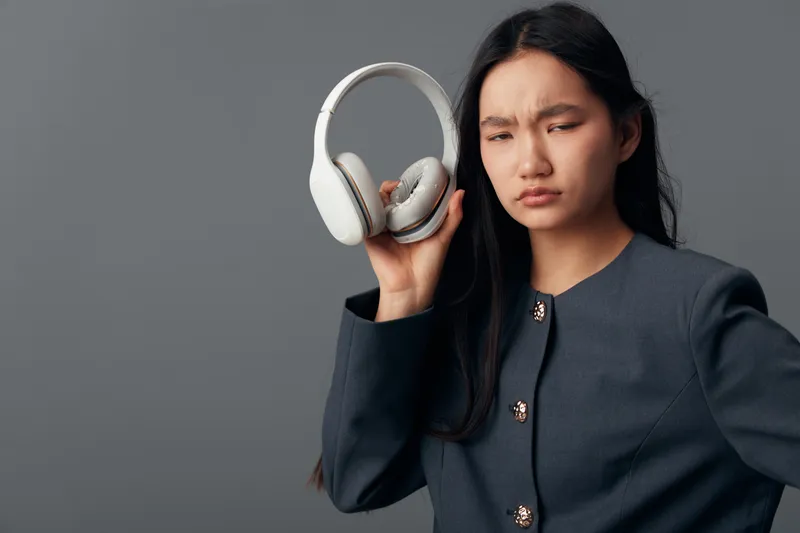 Woman holding wireless headphones with a confused expression on gray background. Female person in dark jacket showing skepticism and uncertainty with modern audio device.