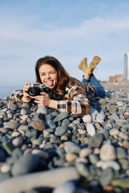 Photographer woman lying on a rocky beach, smiling while using a camera, plaid shirt, sunny outdoor moment among pebbles and shore for creative lifestyle shoot.