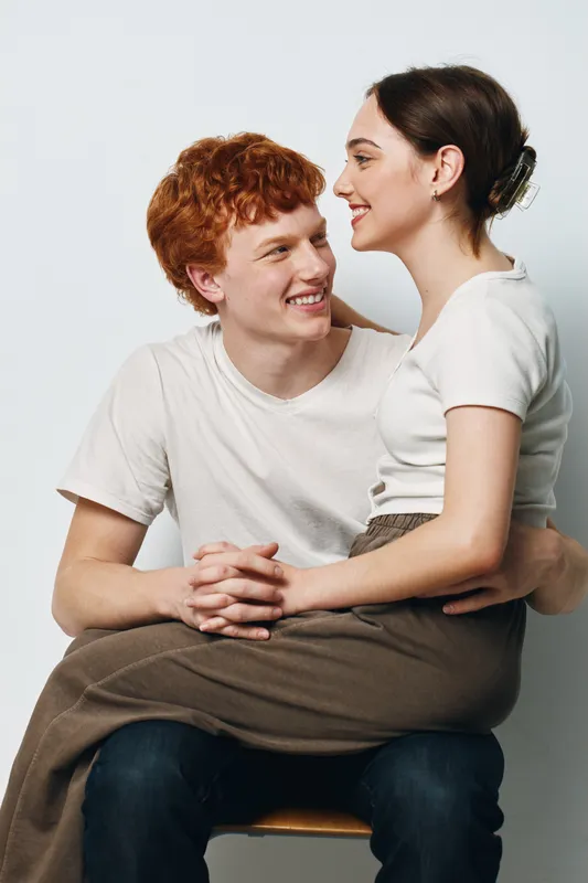 Couple, young man and woman sitting together, smiling and looking at each other, casual clothing, white background, intimacy, happiness and close relationship in studio portrait.