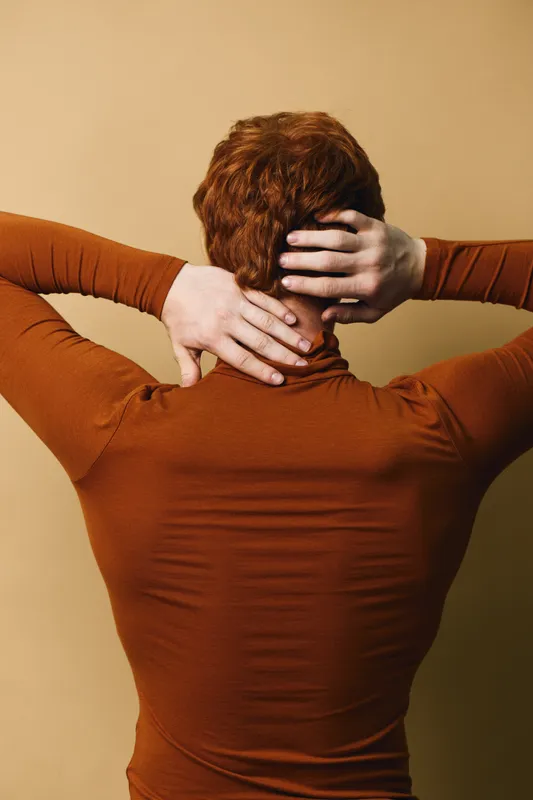 Person with red hair and brown turtleneck facing back and hands on head with beige background for stress or thinking concept.