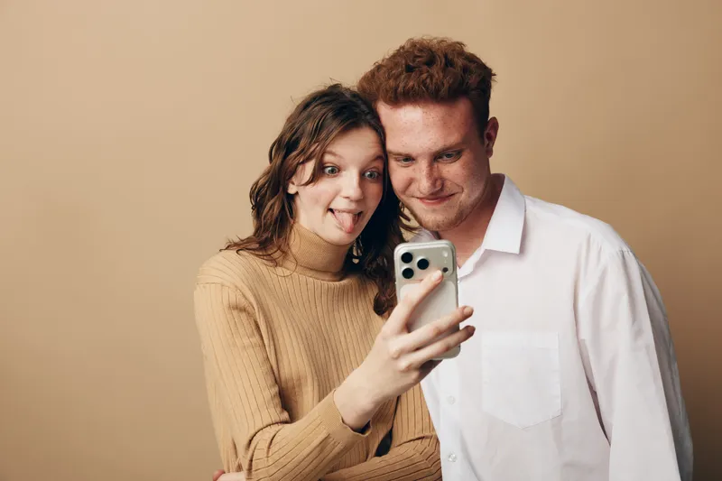 Young couple takes a selfie with a smartphone, smiling and making funny faces. Casual clothing and beige background create a relaxed and playful atmosphere for social media concept.
