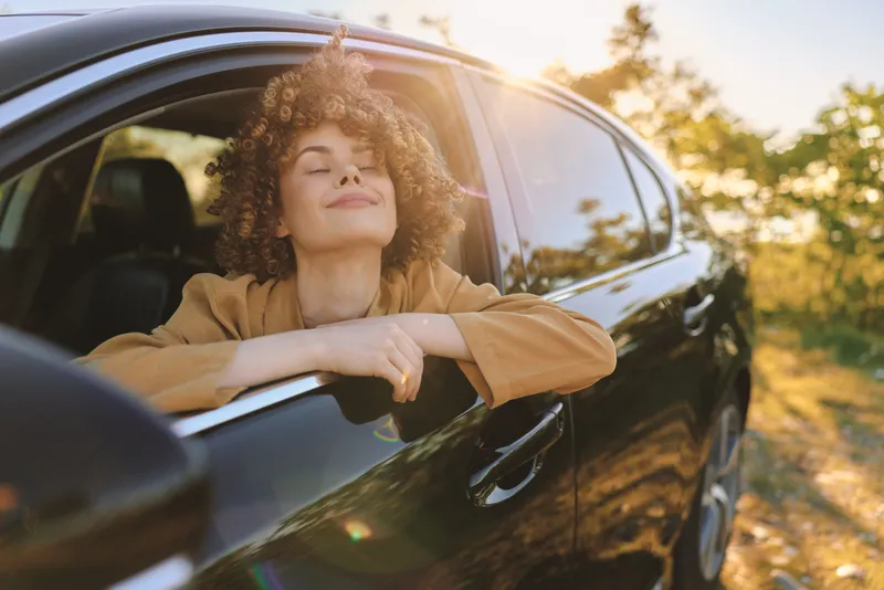 Young woman with curly hair rests her arms on a car window, savoring a carefree sunny afternoon in a retro film color mood, bright light and calm expression.