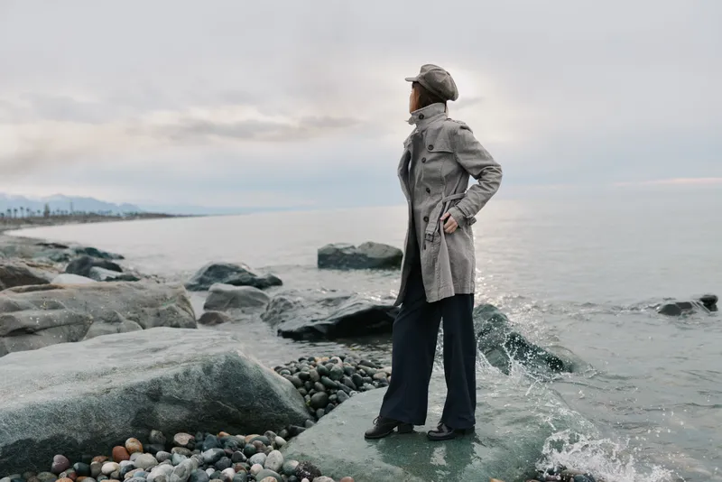 Woman in grey coat and hat stands on rocky shore looking at calm sea under cloudy sky. Concept of contemplation, nature, autumn fashion, and peaceful outdoor moment by water.