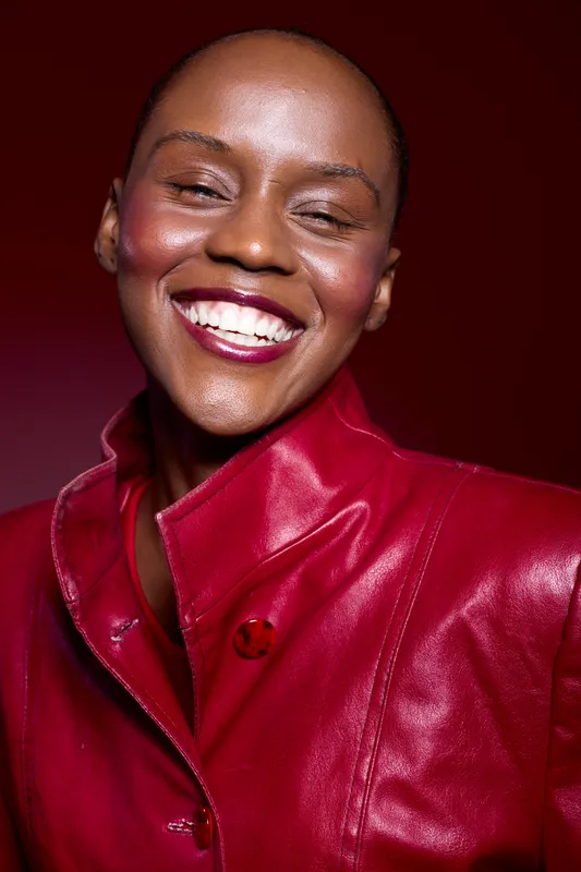 Closeup of happy African woman with dark skin wearing red leather jacket smiling with closed eyes on dark red background. Concept of joy, beauty, fashion, and confident female model.