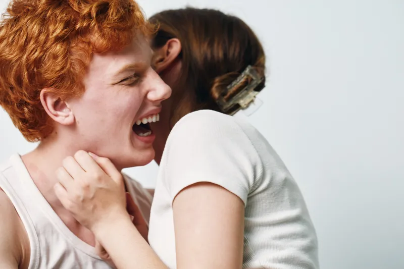 Laughing redhead man and woman whispering secret on white background with casual clothes, happy moment of friendship and close emotional connection indoors.