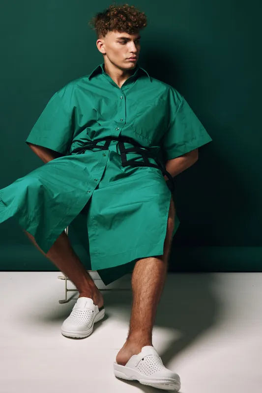 Young man sitting in green oversized shirt dress with black belt and white slip on shoes against dark green background in studio. Fashion model with curly hair and casual style.