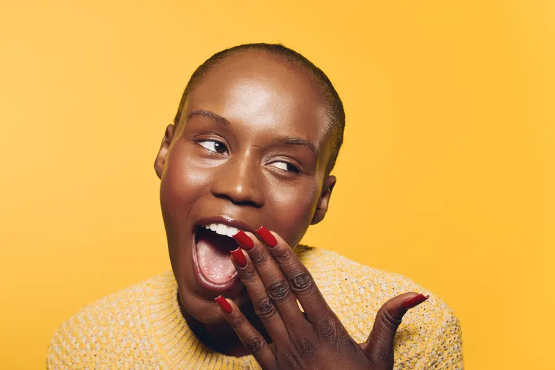Closeup portrait of a happy black woman with dark skin wearing a yellow sweater smiling and covering her mouth with red painted nails on a yellow background. Concept of joy and beauty.
