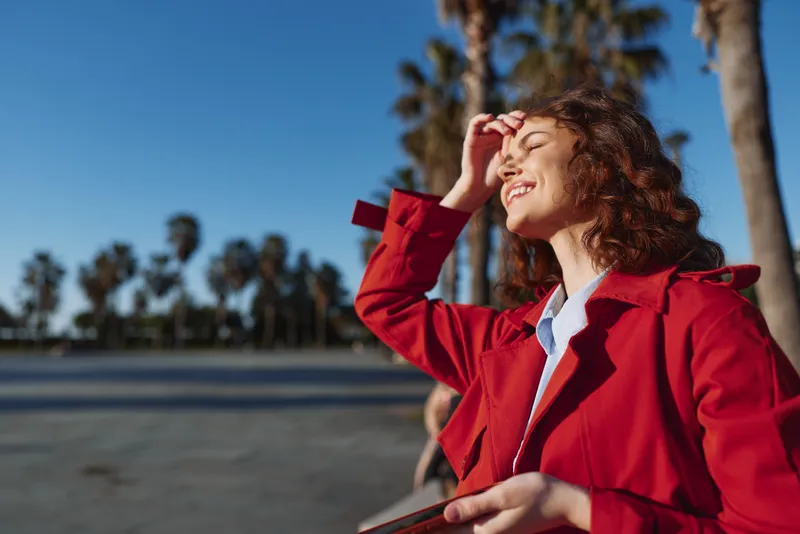 Woman in a bright red coat stands outdoors on a sunny day, shielding her eyes with a smile against palm trees, conveying confidence and fashion.