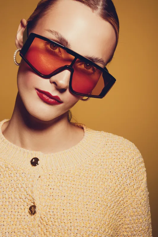 Stylish woman with oversized orange sunglasses and red lipstick poses against warm beige background. Fashion portrait with trendy accessories and confident expression.