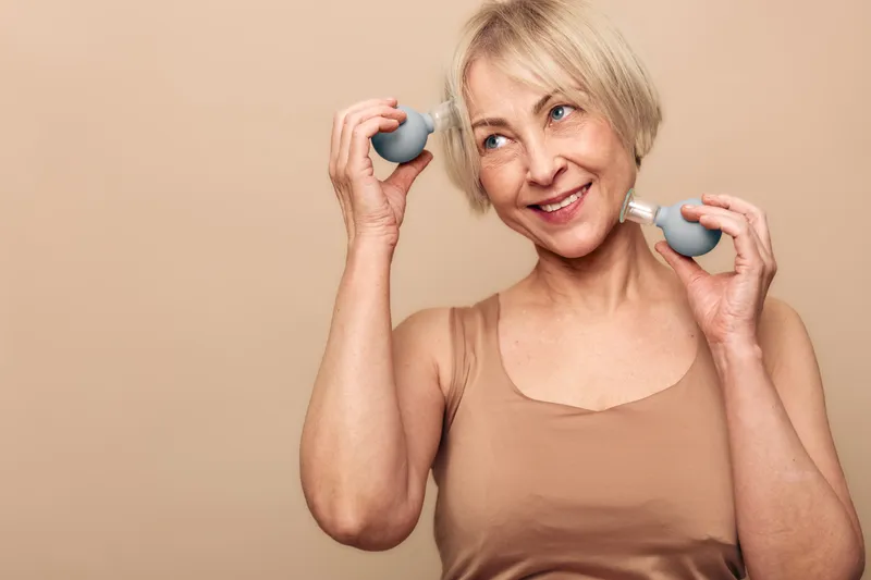 Smiling middle aged woman with short blonde hair holds two gray silicone facial cleansing brushes, demonstrating skincare and beauty routine on beige background.
