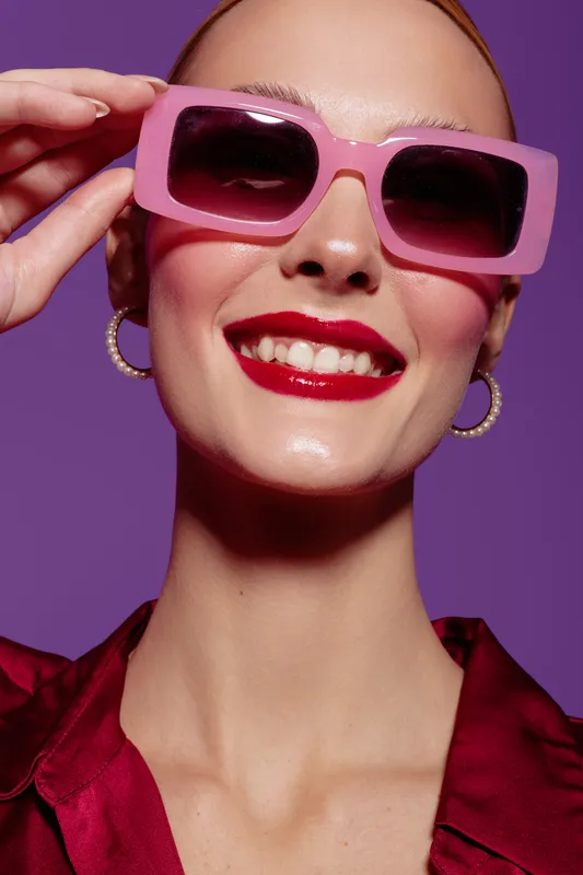 Smiling woman with pink sunglasses and red lipstick poses against purple background. Fashion portrait with bright makeup and joyful expression. Concept of style and beauty.
