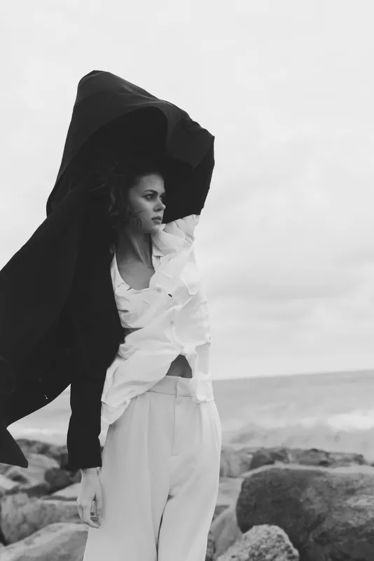 Woman in flowing white outfit on a windy shore, oversized black cloak sweeping behind, dramatic outdoor fashion scene with coastal rocks