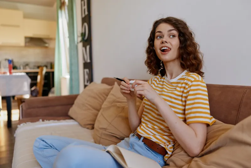 Cheerful woman indoors enjoying a casual home moment, seated on a couch with a game controller, relaxed posture, warm lighting, cozy living space, natural colors, candid lifestyle scene