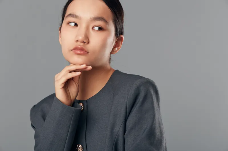 Young woman with thoughtful expression touching chin on gray background, wearing dark stylish sweater, looking sideways for portrait or fashion concept in studio.