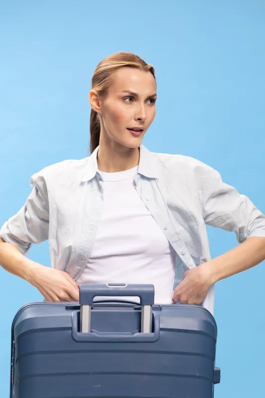 Young woman with light brown hair stands behind blue suitcase wearing white shirt on blue background. Concept of travel, packing, tourism, preparation, and vacation planning.