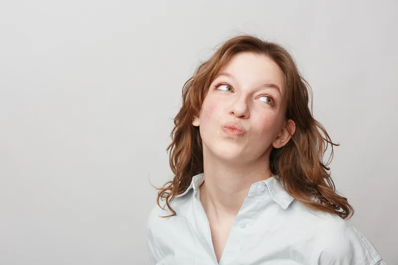 Young woman with curly hair looks up thoughtfully with pursed lips against a plain light background. Concept for thinking, curiosity, and contemplation.