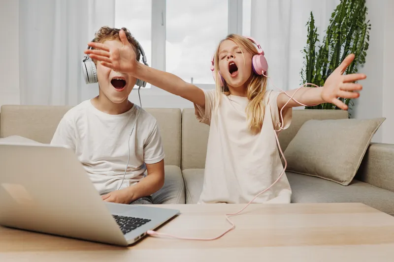 Two children with headphones enjoy music at home, sitting on a sofa with laptop in front. The girl sings passionately while the boy smiles, sharing joyful moments together.