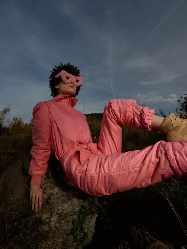 Puppet wearing a pink suit and sunglasses lounges on a rocky outcrop in an open field, dramatic sky above, capturing surreal fashion and playful attitude in a natural outdoor setting.