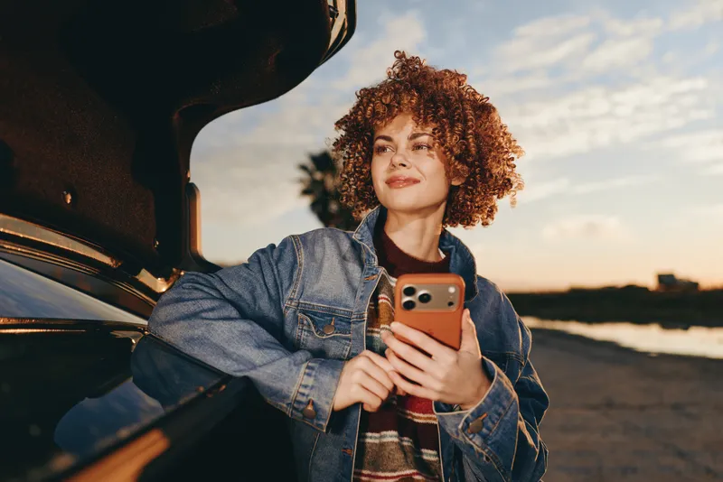 Curly haired woman lifestyle near car door, holding smartphone, denim jacket, smiling at golden hour sunset on road, casual travel vibe, outdoors close view, soft light moment
