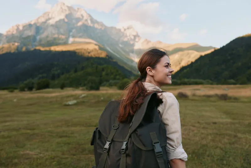 Cheerful woman outdoors, smiling over her shoulder, hiking with a backpack, mountains and valley in warm sunlit light, natural lifestyle moment